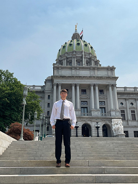 A young man in a white shirt and tie stands on the steps of the Pennsylvania State Capitol, with its green dome in the background.